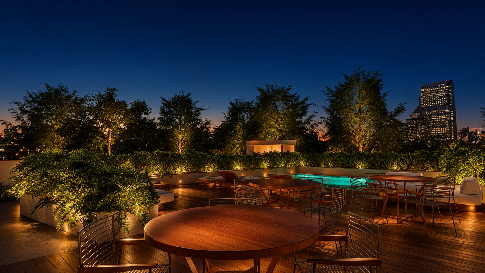 Lounge do rooftop com mesas, paisagismo e piscina ao fundo.